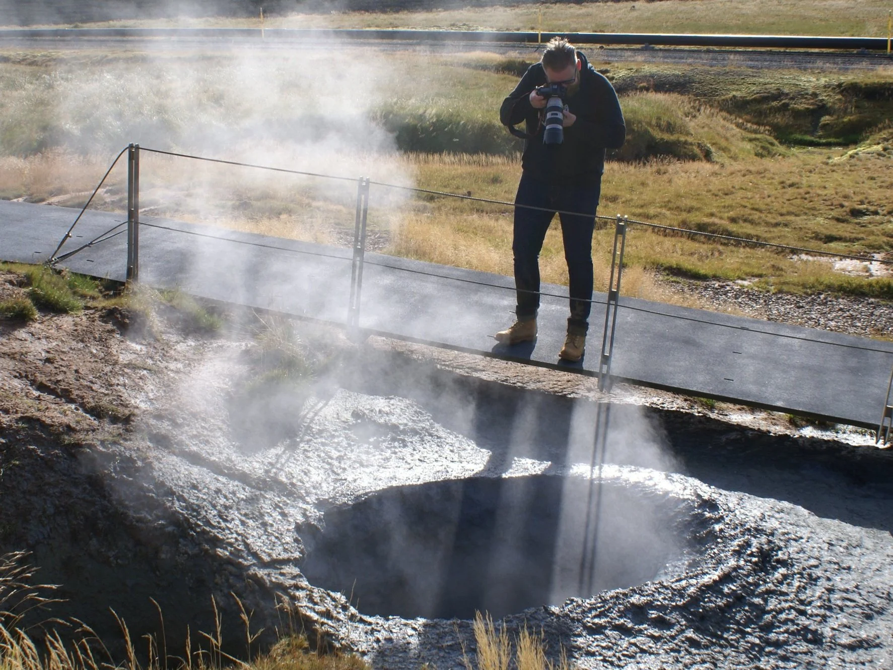 A photographer on a designated walkway beside a steaming hot spring at Hveradalir